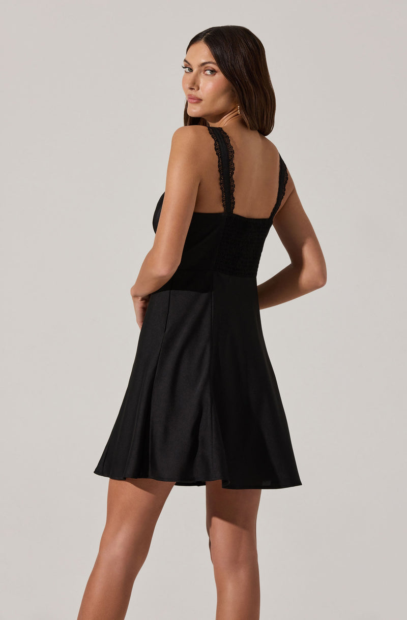 A woman is wearing a black sleeveless dress with wide shoulder straps and lace detailing along the straps. The dress has a fitted bodice and a flared skirt that falls above the knees. The fabric appears smooth and slightly shiny, giving it an elegant look.
