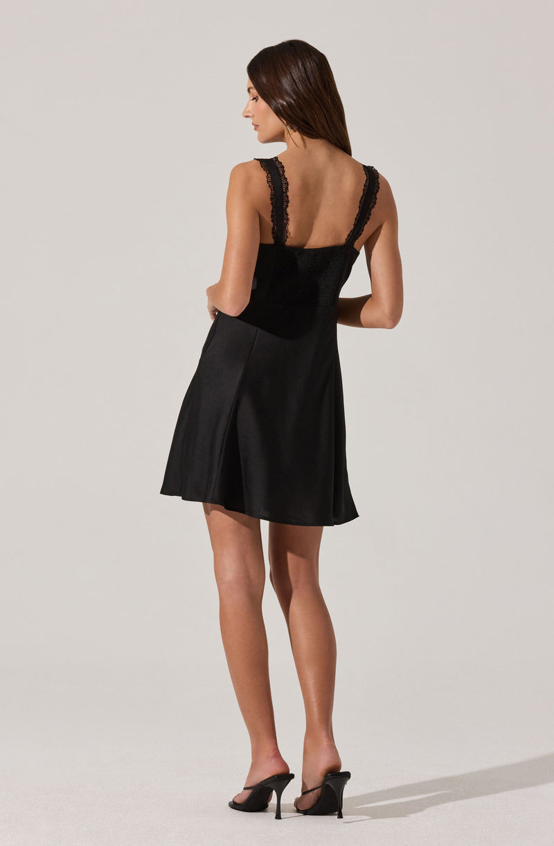 A woman is wearing a black sleeveless dress with lace straps on the shoulders. The dress has a fitted bodice and a flared skirt that falls above the knees. She is also wearing black high-heeled sandals with thin straps.