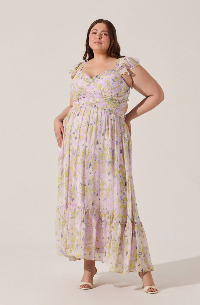 A woman is wearing a long, flowy maxi dress with a pastel floral pattern in shades of pink, yellow, purple, and green. The dress features ruffled shoulder straps, a fitted bodice with a ruched design, and a tiered skirt that reaches the floor. She pairs it with beige open-toe heels.