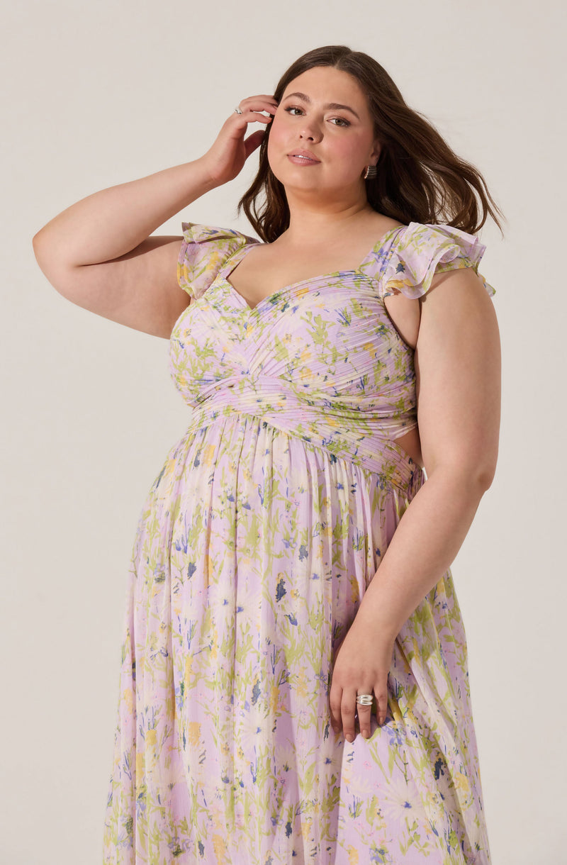 A woman is wearing a pastel-colored, floral-patterned dress with ruffled sleeves, a sweetheart neckline, and a flowing skirt. The dress features light pink, yellow, green, and purple hues with a delicate, slightly textured fabric. She has her right hand gently touching her hair and her left hand resting by her side, showcasing a silver ring.