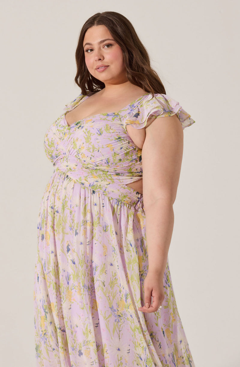 A woman wearing a flowy, pastel-colored dress with a floral pattern in shades of pink, yellow, green, and purple. The dress features ruffled cap sleeves and a fitted bodice with horizontal pleats. The skirt is long and gathered, creating a soft, flowing silhouette.
