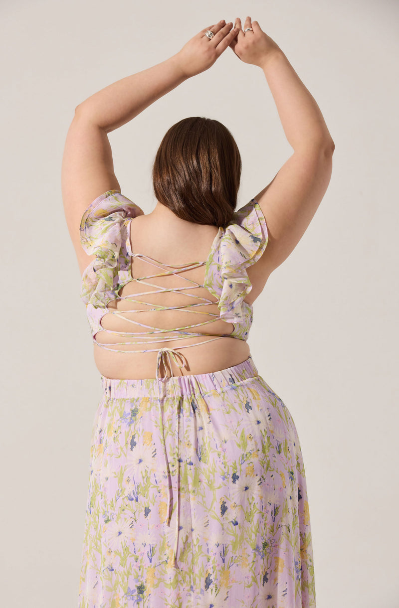 A person with short brown hair is wearing a pastel-colored, floral-patterned outfit. The top features ruffled sleeves and a back design with crisscrossing ties, revealing part of the back. The matching skirt has an elastic waistband with a drawstring and a light, flowy fabric adorned with a floral print in shades of pink, purple, yellow, and green.