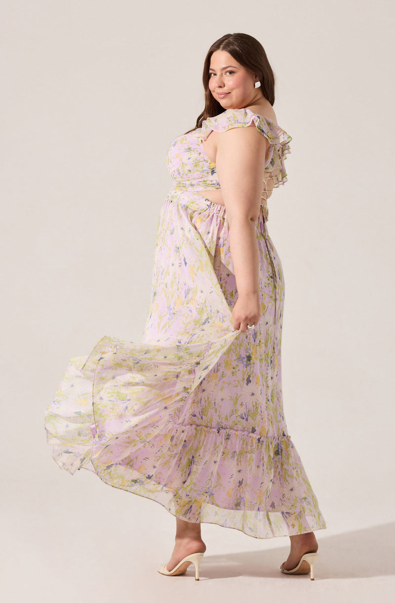 A woman is wearing a light, flowy, floral patterned dress with pastel shades of pink, yellow, and lavender. The dress features ruffled straps and a cut-out detail at the waist. She is holding the hem of her dress, which has a ruffled hemline, and is standing in high-heeled shoes.
