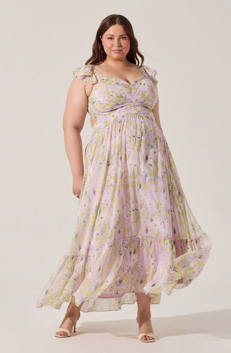 A woman is wearing a long, flowy dress with a pastel floral pattern consisting of light pink, yellow, purple, and green colors. The dress has short, ruffled sleeves and a fitted bodice with gathered fabric at the bust, creating a ruched effect. The skirt is tiered, with a ruffle at the bottom hem, and the fabric appears lightweight and airy. She is also wearing open-toed, beige high heel shoes.