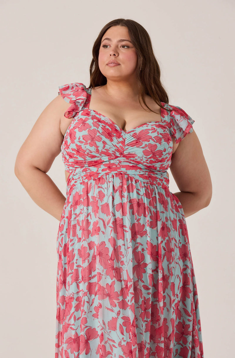 A woman is wearing a sleeveless dress with ruffled shoulder straps. The dress features a light blue background with a red floral print, gathered at the bust with a ruched detail, and flows down to a long, gathered skirt.