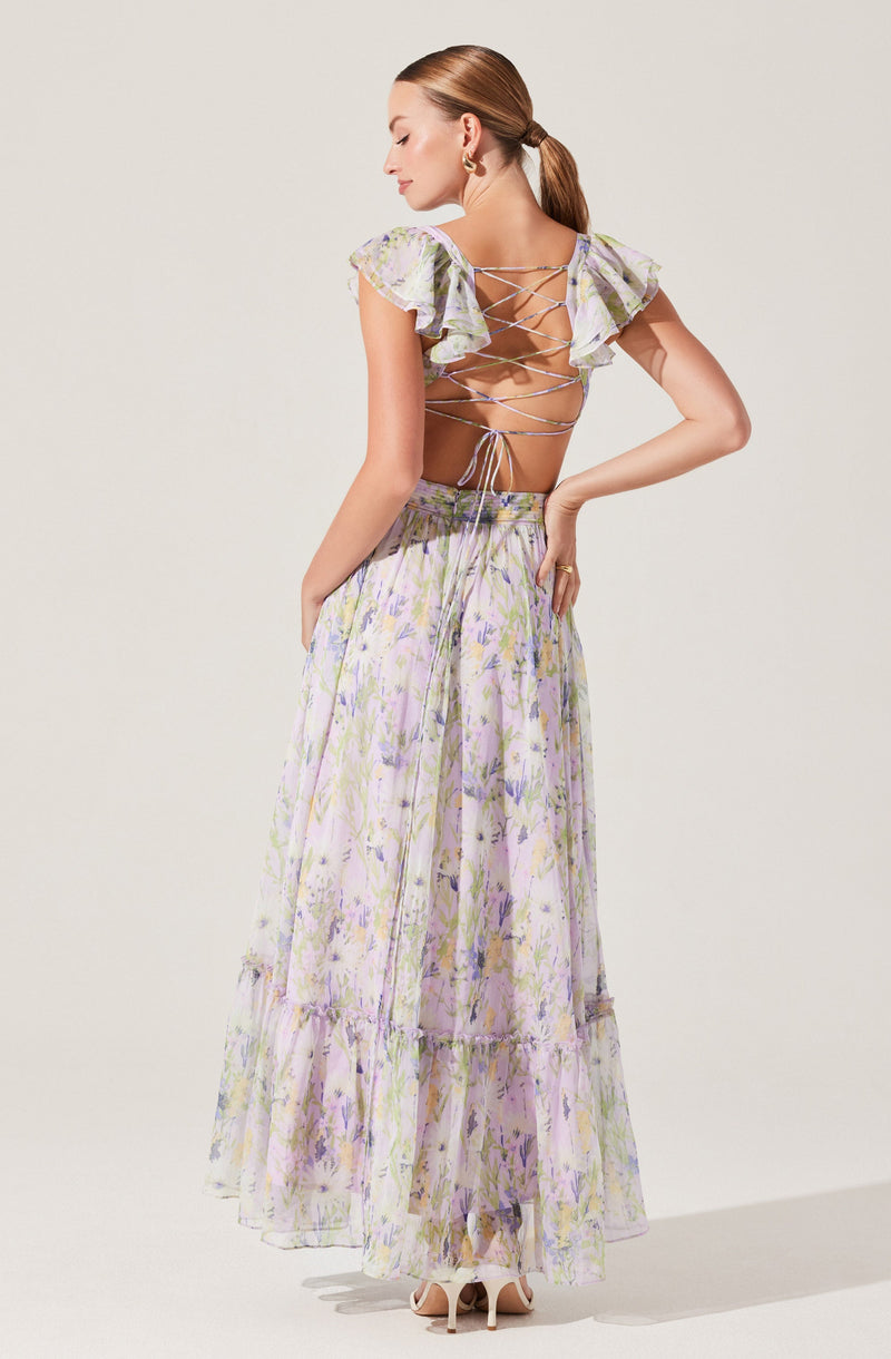 A woman wearing a two-piece outfit with a pastel floral pattern. The top features ruffled short sleeves and a back laced-up design with crisscrossing ties. The long, flowing skirt also displays a matching floral pattern and has a tiered ruffle detail near the hem. She is wearing high-heeled shoes and has pulled her hair back into a ponytail.