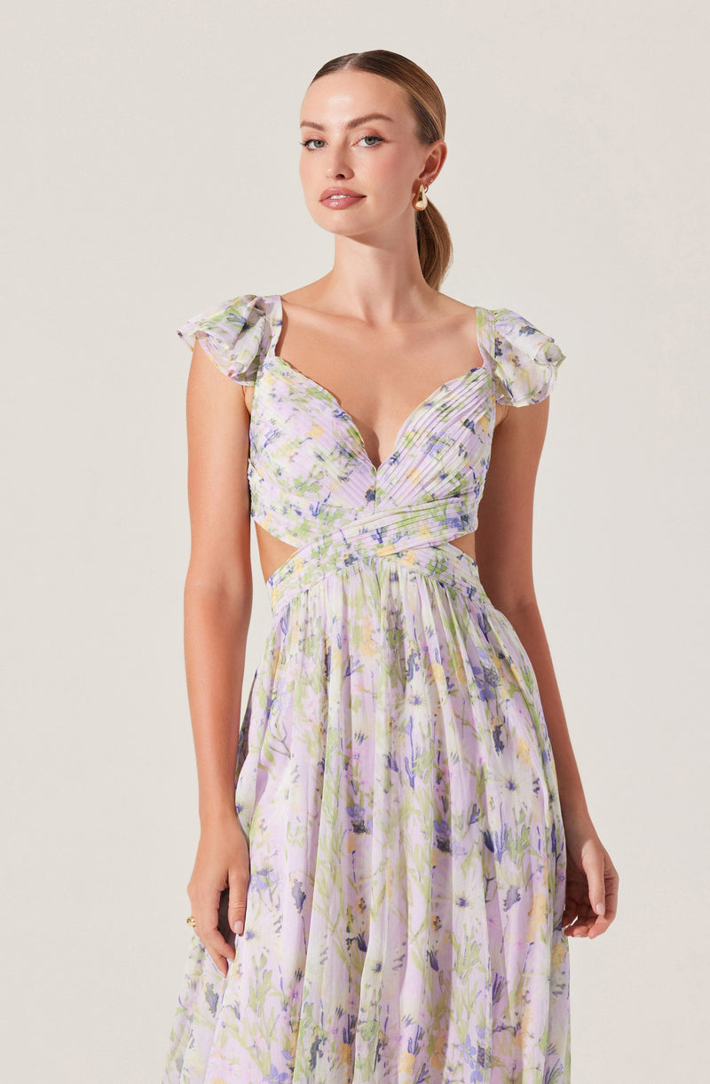 A woman wearing a long, flowy dress with a floral pattern in pastel colors including purple, green, and yellow. The dress has a deep V neckline, pleated bodice, and short puff sleeves with ruffle details. The fabric appears lightweight and breathable.