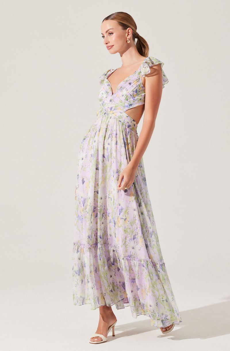 A woman is wearing a long, flowing dress with a pastel floral pattern. The dress features ruffled shoulder straps, a deep V-neckline, and cutout sides at the waist. The fabric appears light and airy, with a tiered skirt that reaches the floor. She is also wearing open-toed high heels.