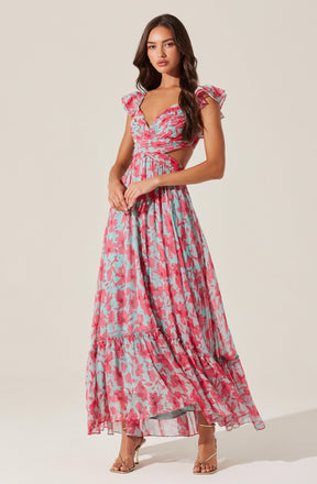 A woman is wearing a long, colorful dress with a pink, red, and light blue floral pattern. The dress features short, ruffled sleeves, a sweetheart neckline, cutouts at the waist, and a flowing, tiered skirt. She is also wearing strappy, high-heeled sandals.