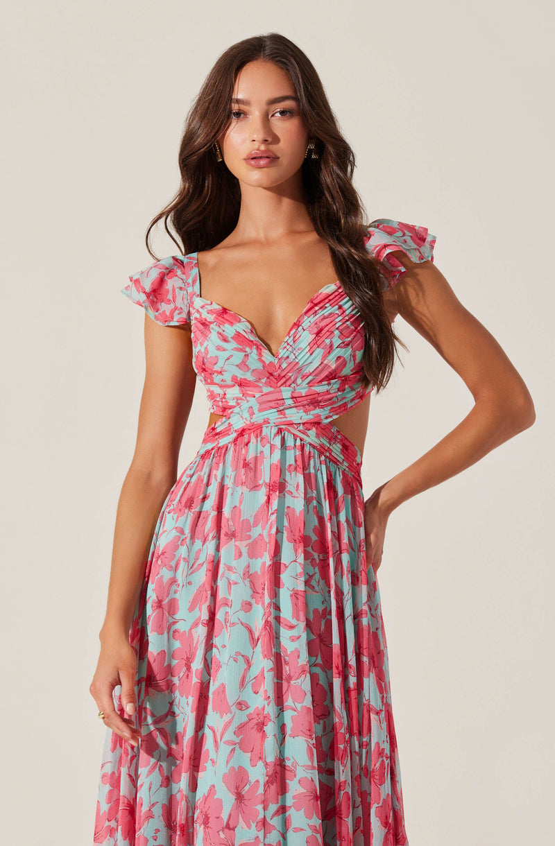 A woman is wearing a long, flowing dress with a floral pattern in pink and light blue. The dress features ruffled short sleeves, a ruched bodice with a sweetheart neckline, and a waistline that accentuates her figure.