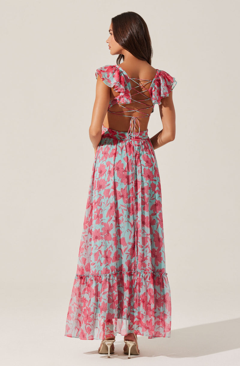 A woman wearing a two-piece outfit with a floral pattern. The top has ruffled sleeves and is laced up at the back with crisscrossing ties. The long, flowy skirt features the same floral pattern and has a tiered ruffle near the hem. She is wearing high-heeled shoes and standing against a plain background.