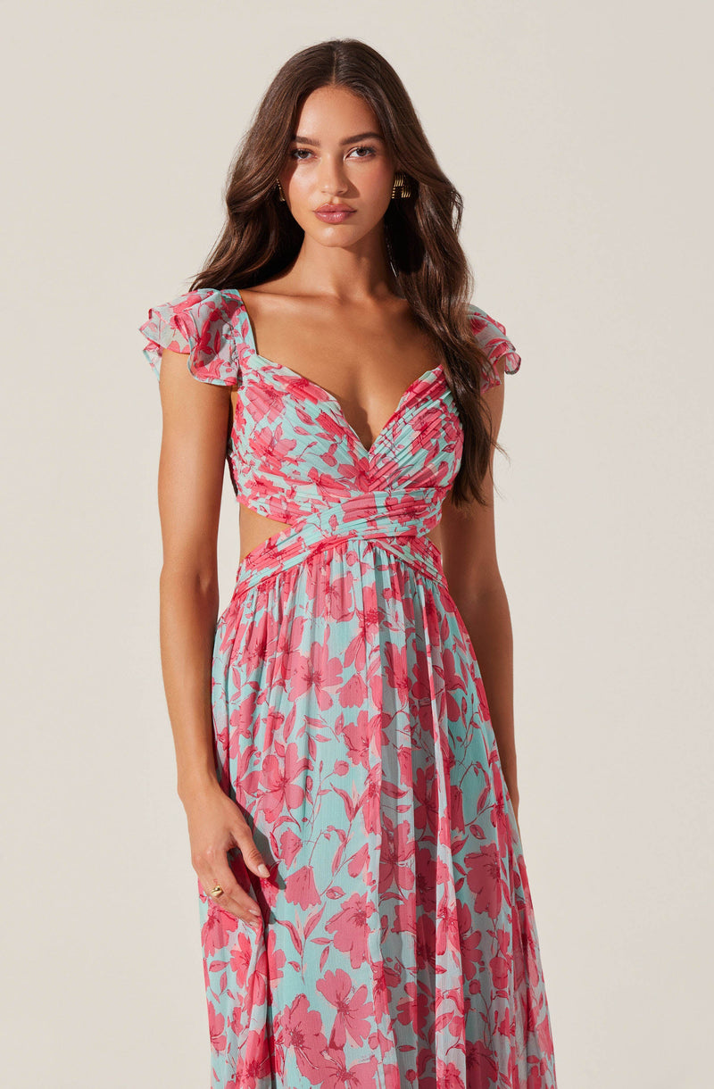 A woman is wearing a long, flowy dress with a floral print. The dress has a fitted bodice with a sweetheart neckline, short ruffled sleeves, and cut-out sides at the waist. The fabric features a pattern of pink flowers on a light blue background.