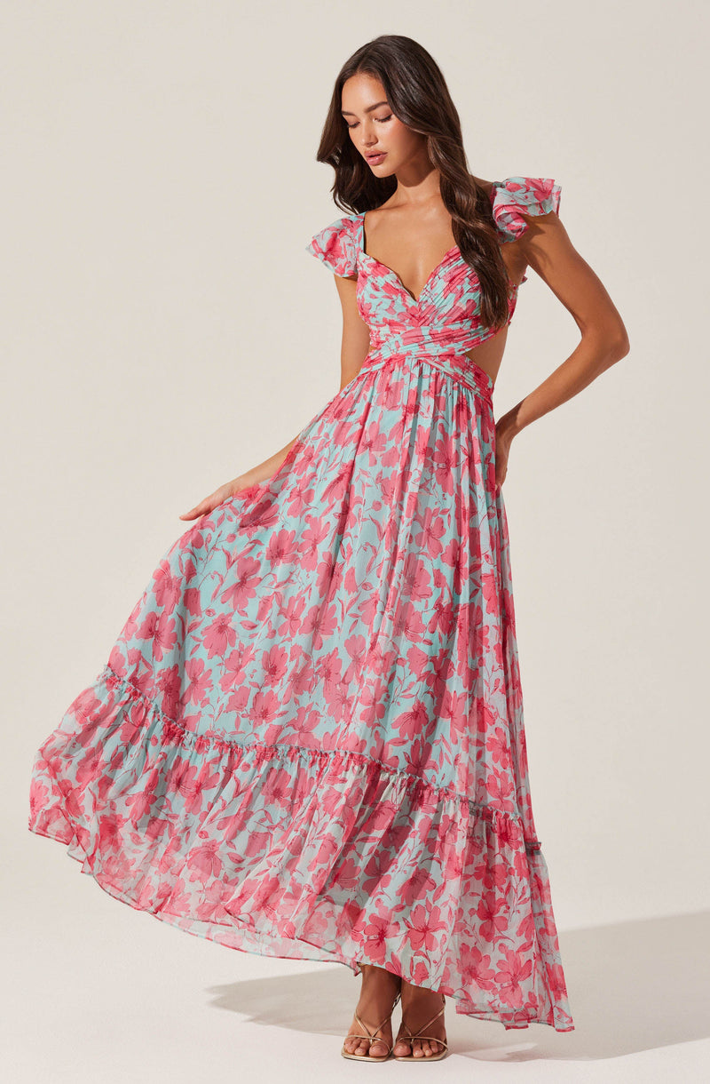 A woman is wearing a long, flowing dress with a pink and light blue floral pattern. The dress features ruffled cap sleeves, a fitted bodice with a V-neckline, and cut-out sides at the waist. The skirt has a tiered design with ruffles at the hem, creating a breezy, summery look.