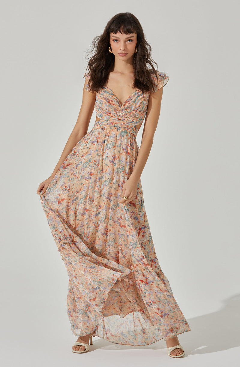 A woman is wearing a long, flowy dress with a floral pattern in shades of peach, blue, and purple. The dress has a V-neckline, short flutter sleeves, and a fitted waist that accentuates her figure. She is also wearing white open-toe heels with ankle straps.