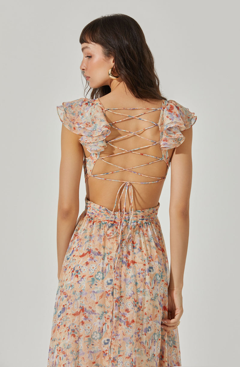 A woman is wearing a backless dress with short, ruffled sleeves and a floral pattern in soft pastel colors. The dress features crisscrossing laces on the back that tie at the bottom, revealing her back and shoulders. The skirt of the dress is high-waisted and flows down, matching the floral pattern and colors of the top.