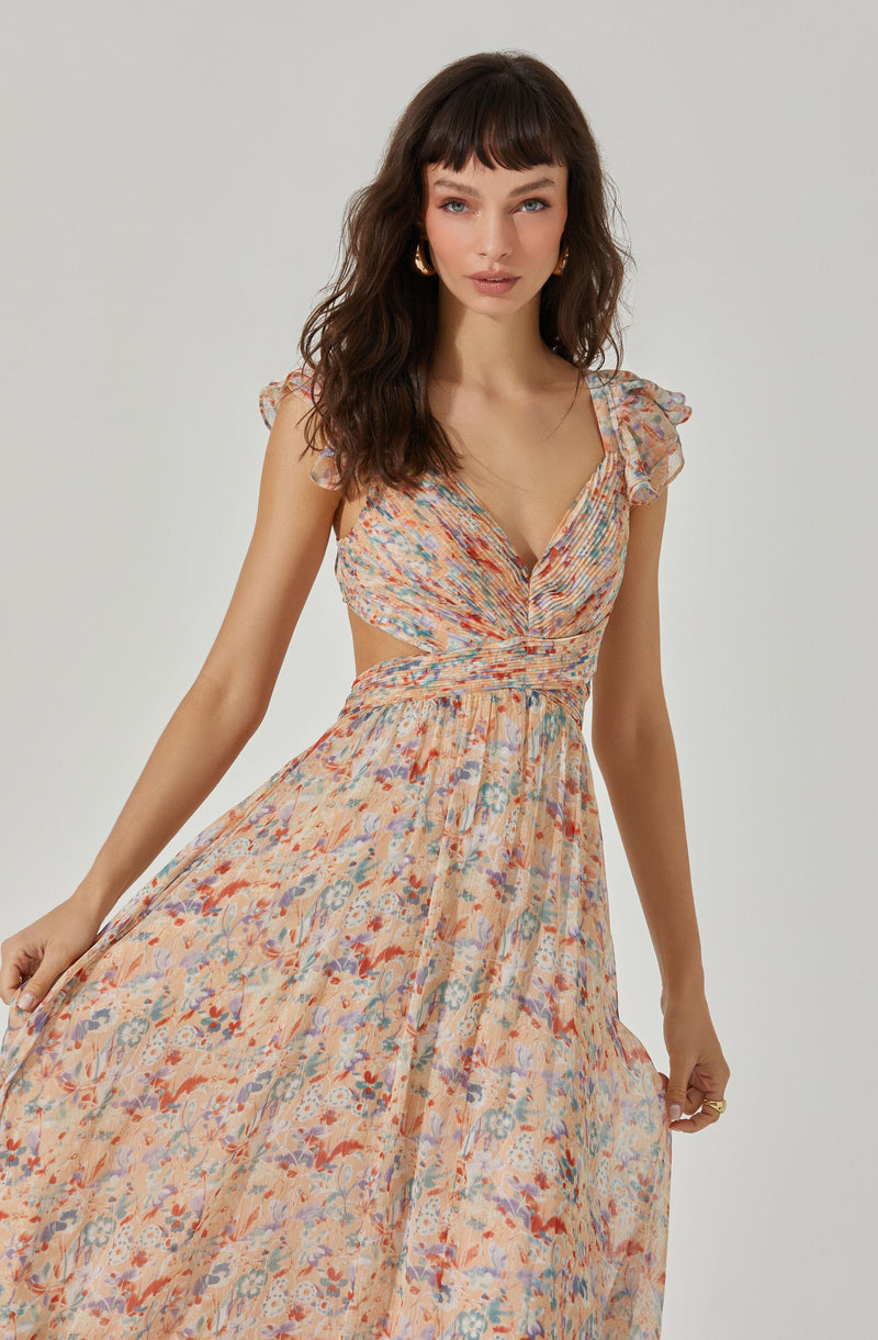 A woman is wearing a flowy, floral-patterned dress in shades of peach, blue, and red. The dress features ruffled shoulder details, a deep V neckline, and a smocked waistline that accentuates her figure.