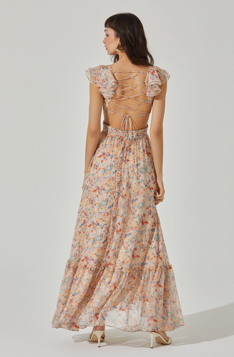 A woman is wearing a long, flowing dress with a floral pattern in pastel shades of pink, peach, blue, and cream. The dress features ruffled cap sleeves, an open back with crisscross laces, and a high waistline. The skirt is gathered at the waist and has a tiered, ruffled hem. She is also wearing white high-heeled shoes.