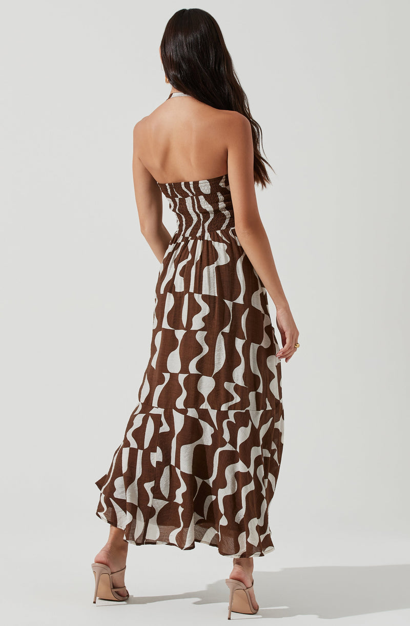 A woman is wearing a strapless, maxi dress with a fitted, ruched bodice. The dress features a bold, abstract pattern in shades of brown and beige, with a flowy, tiered skirt that reaches the floor. She is also wearing high-heeled sandals in a neutral tone.