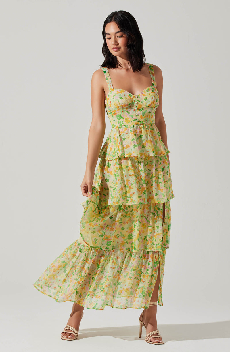A woman is wearing a long, flowy dress with a colorful floral pattern. The dress has a fitted bodice with buttons down the front, wide shoulder straps, a peplum waist, and tiered, ruffled layers that extend to the floor. She is wearing open-toed high-heeled sandals in a light, neutral color.