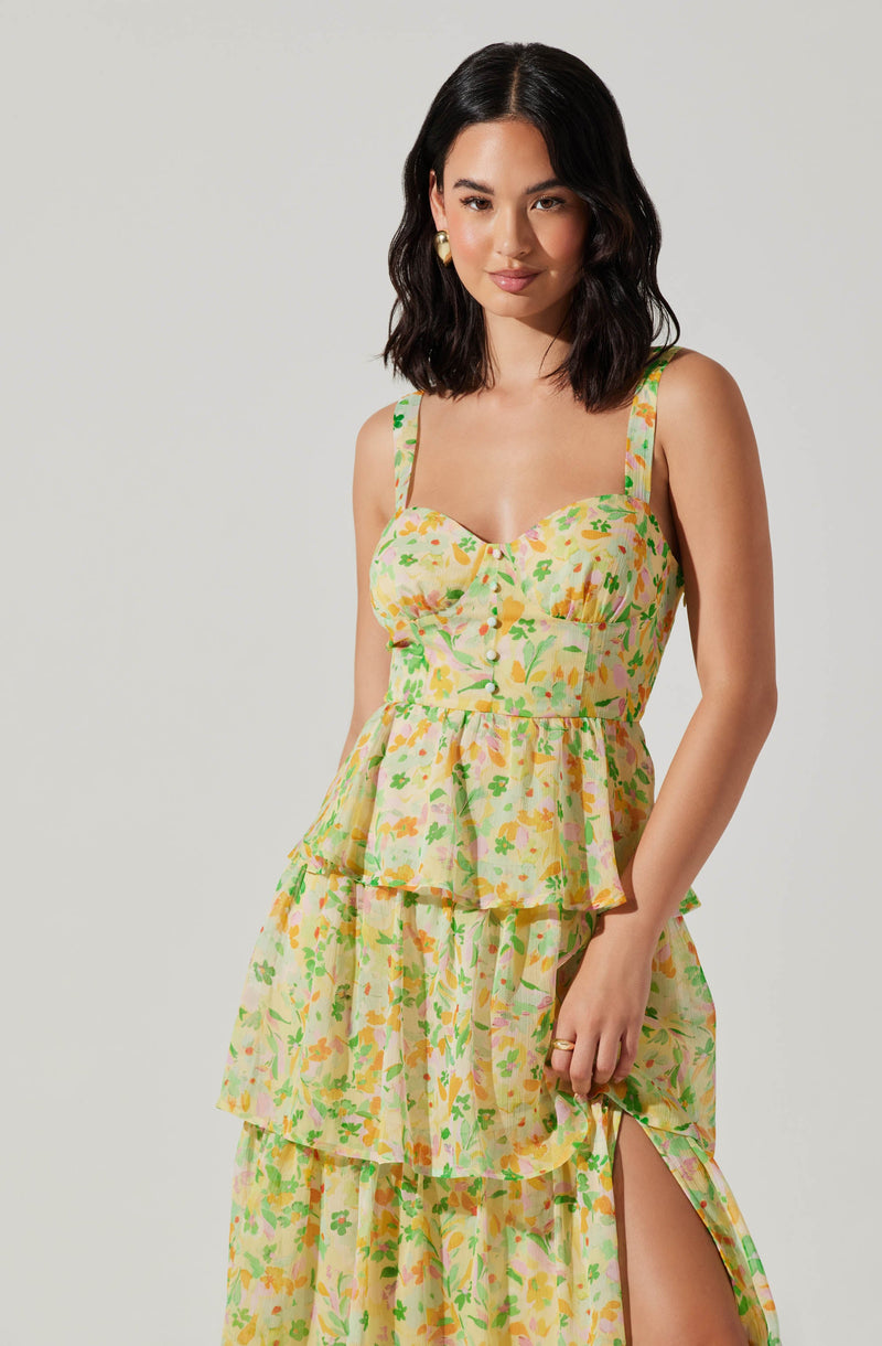 A woman wearing a colorful, sleeveless, floral dress with a fitted bodice, button details down the front, and a tiered, flowing skirt with a slit on one side. The dress features pastel shades of yellow, green, pink, and orange, and has wide shoulder straps. She also has gold hoop earrings.