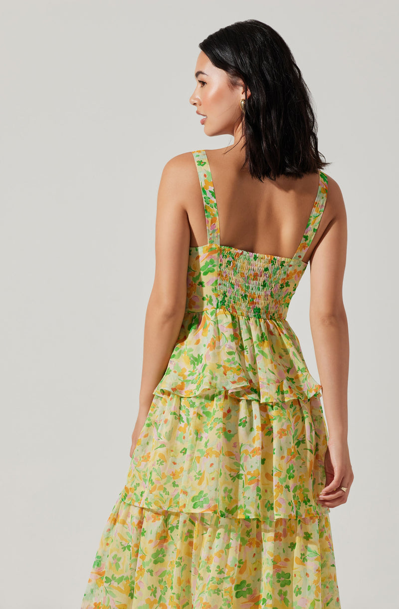 A woman is wearing a sleeveless dress with wide shoulder straps. The dress features a colorful floral pattern with shades of yellow, green, pink, and orange. The bodice is smocked, and the skirt has layered ruffles. The fabric appears lightweight and flowing.