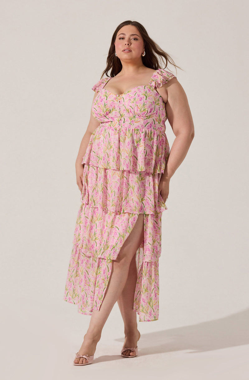 A woman is wearing a sleeveless, pink floral dress with ruffled shoulder straps. The dress has a fitted bodice, a layered skirt with a high slit on the left side, and features green and yellow leaf patterns throughout. She is also wearing open-toe heels.