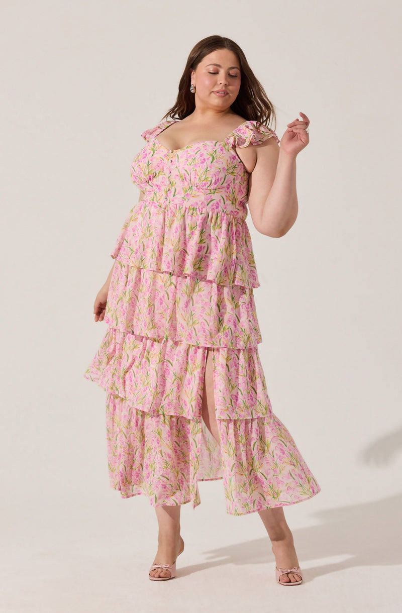 A woman is wearing a pink, floral-patterned dress with green and yellow accents. The dress features a fitted bodice with ruffled shoulder straps and an A-line skirt with multiple tiers of ruffles, creating a layered look. She is also wearing nude-colored open-toe heels.