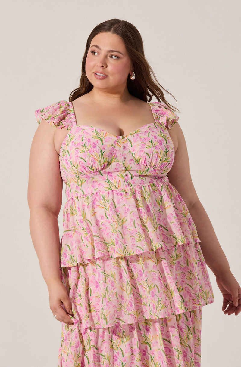 A woman with long, wavy brown hair is wearing a pink floral dress with green and yellow accents. The dress features ruffled cap sleeves and a sweetheart neckline. It has a tiered, flowing skirt with multiple layers, creating a bohemian, romantic style.