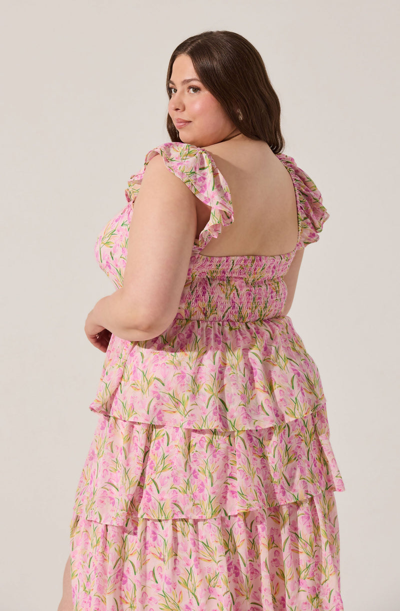 A woman is wearing a pink floral dress with a square neckline that has a smocked bodice. The dress features layered ruffles with a delicate green and pink floral pattern. The sleeves are short and ruffled, adding a feminine touch to the outfit.