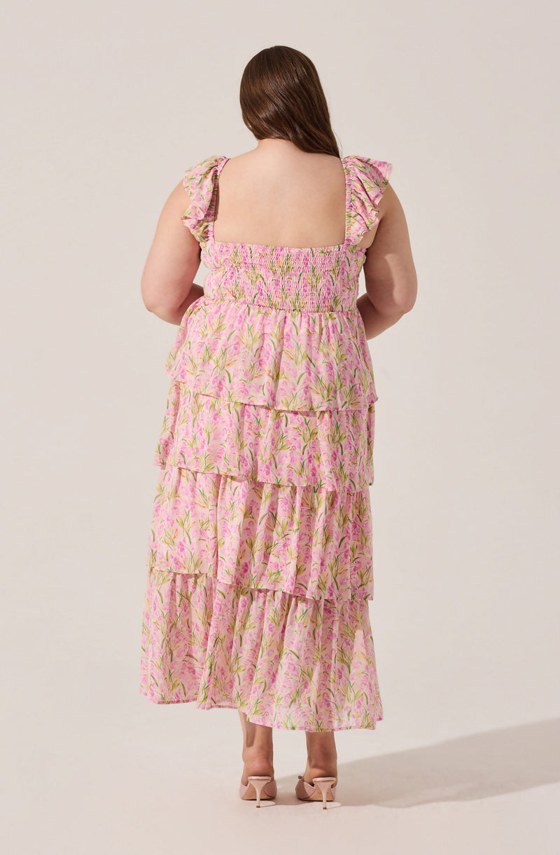 A woman is wearing a long, flowy dress with multiple tiers. The dress features a pink and green floral pattern, with ruffled shoulder straps and a gathered, elasticized neckline. The fabric appears lightweight and airy.
