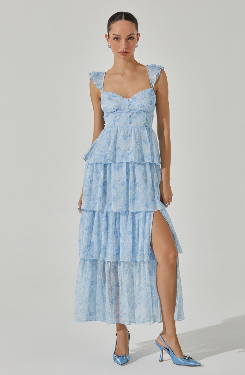 The woman is wearing a light blue, tiered maxi dress with a floral pattern. The dress features wide shoulder straps with ruffled details, a fitted bodice with a sweetheart neckline, and multiple layered ruffles that create a flowing silhouette. One side of the skirt has a high slit revealing her leg. She completes the look with matching light blue high-heeled shoes with bows on the front.