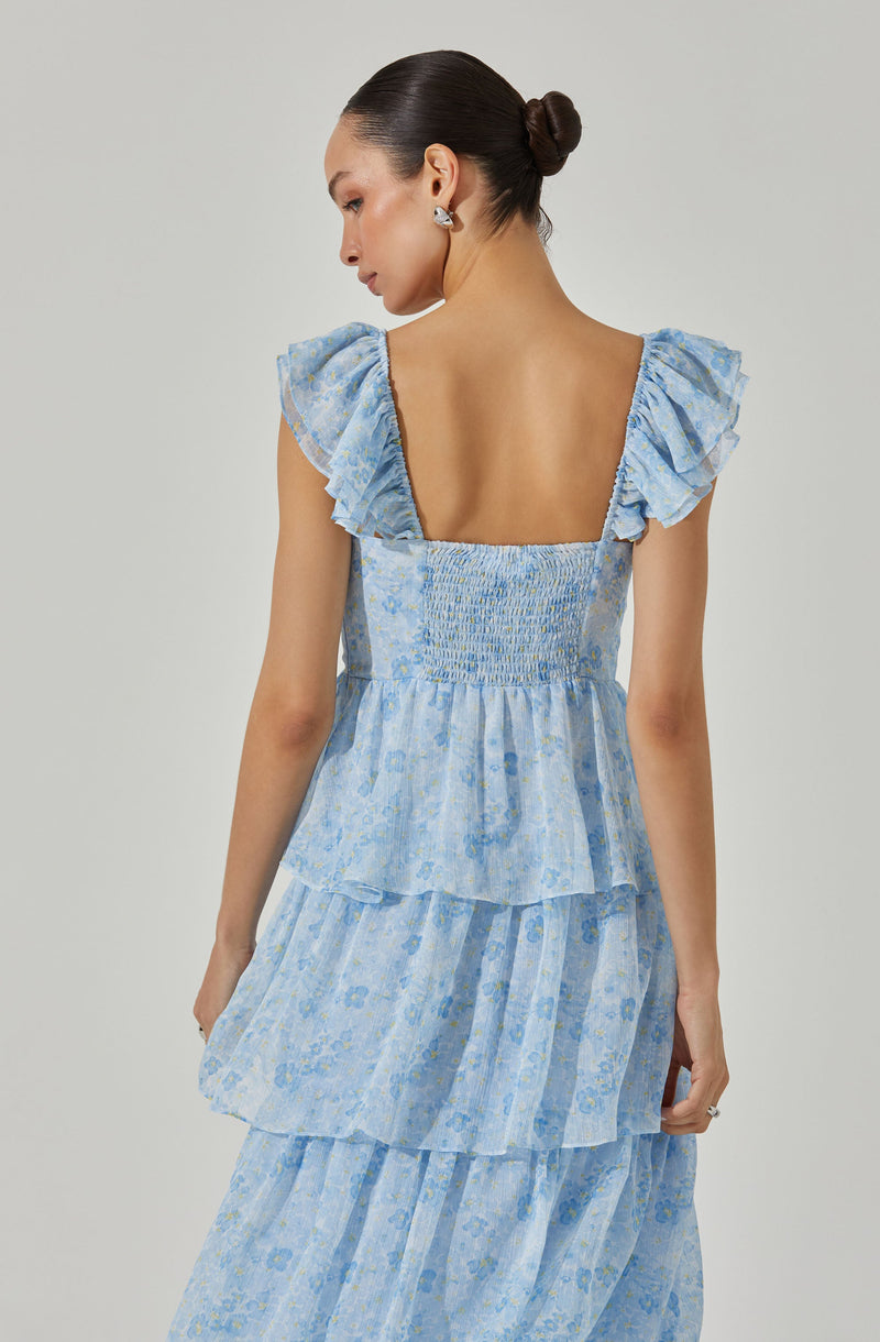 A woman is wearing a light blue, floral-printed dress with ruffled cap sleeves. The dress features smocked detailing at the bodice and multiple tiered layers in a flowing style. The fabric has a subtle floral pattern in shades of blue and yellow.