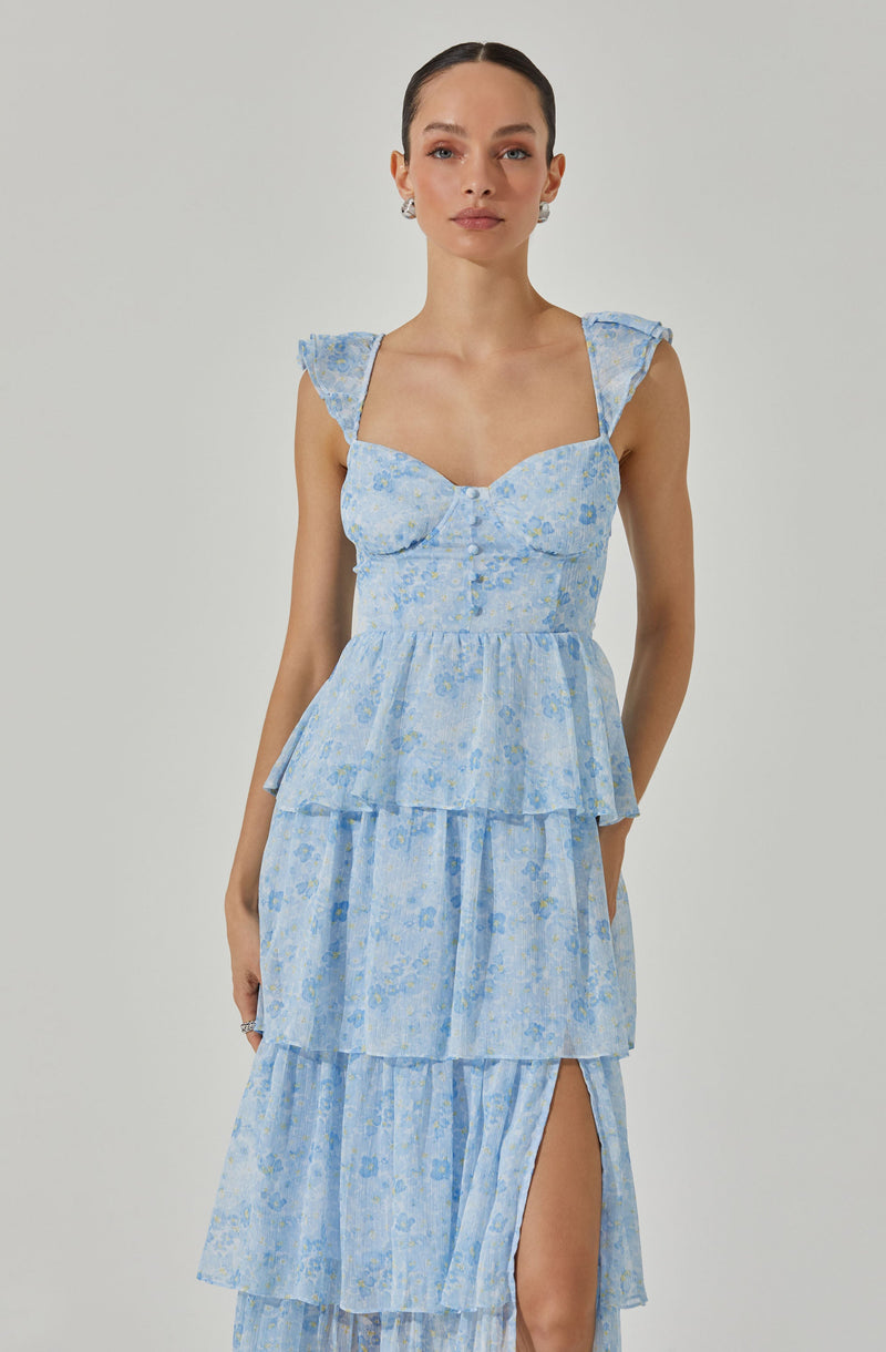 The dress is a light blue, tiered, floral print garment with a fitted bodice featuring a sweetheart neckline and ruffled shoulder straps. It has multiple ruffled layers cascading down to a high slit on one side.