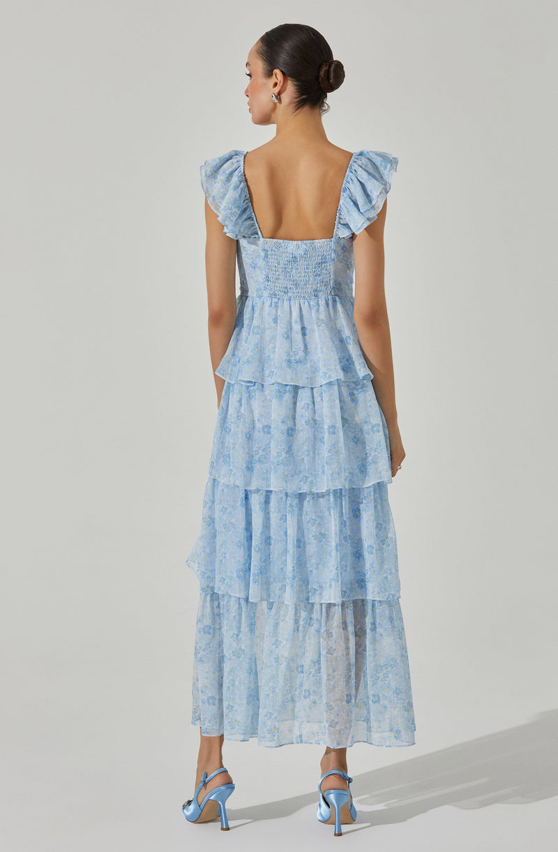 A woman is wearing a light blue, tiered dress with a floral pattern. The dress features smocked fabric at the chest, ruffled cap sleeves, and a deep square neckline at the back. She is also wearing matching light blue high-heeled sandals.