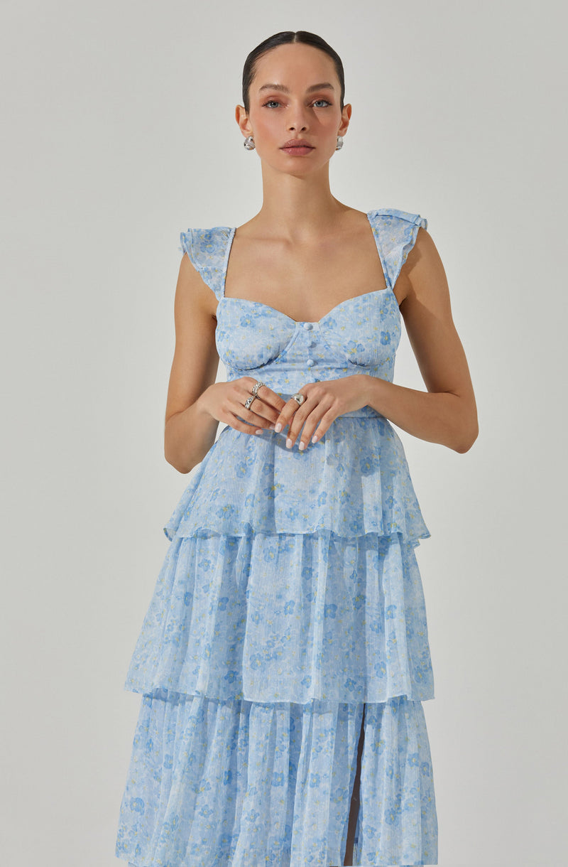 A woman is wearing a light blue, tiered dress with a floral print. The dress has a fitted bodice with a sweetheart neckline and ruffled shoulder straps. The skirt features multiple layers of fabric, creating a flowy and voluminous look.