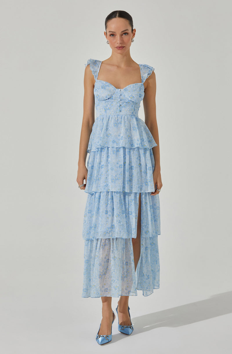 The woman is wearing a light blue, tiered maxi dress with a delicate floral pattern. The dress features ruffled shoulder straps, a fitted bodice with a sweetheart neckline, and multiple layered skirts with a slit showing her right leg. She also has on matching light blue high heels with bow details.