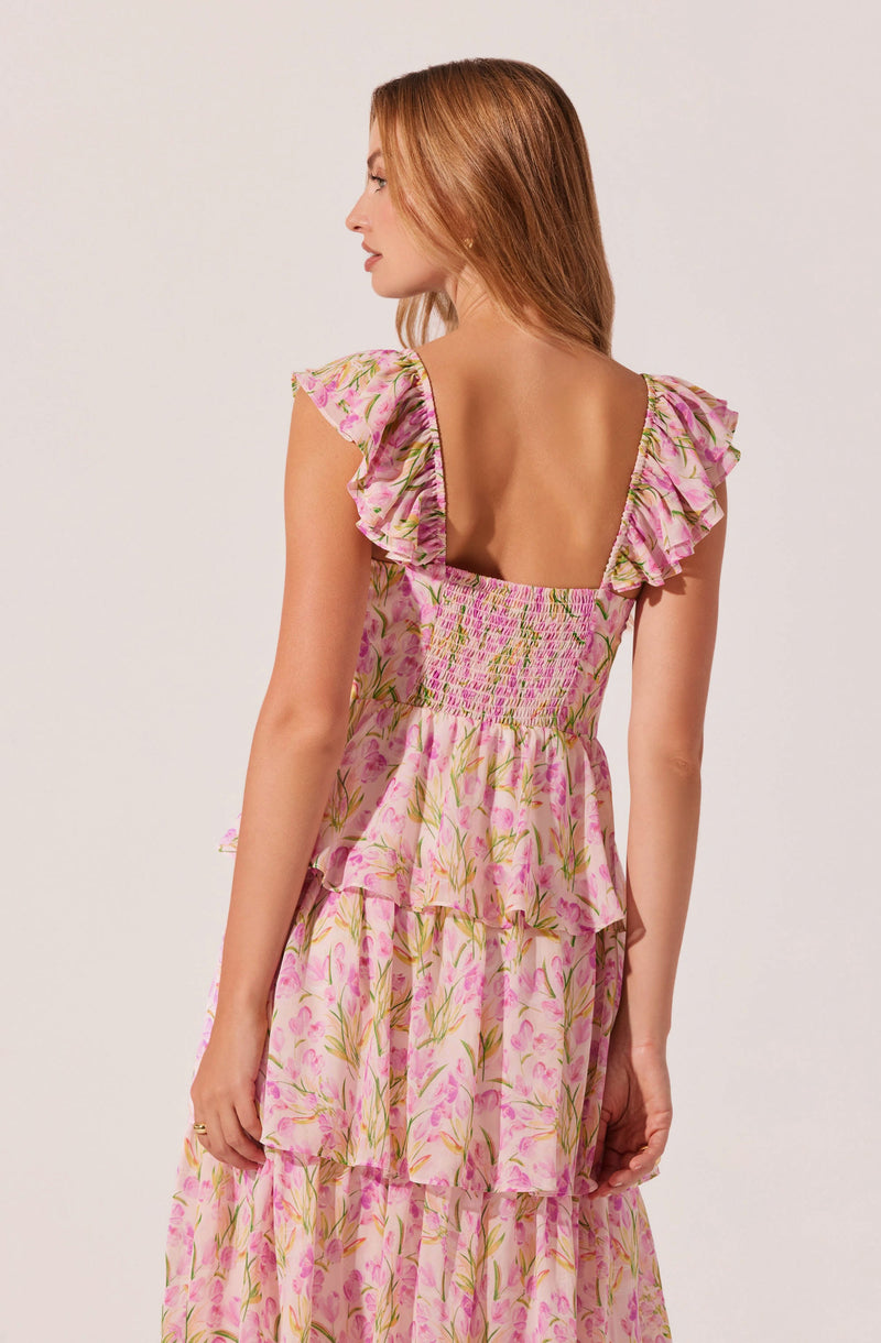 A woman is wearing a pink floral dress with green and yellow accents. The dress features a square neckline and ruffled cap sleeves. The waist is smocked, and the skirt consists of multiple tiered, flowing layers.