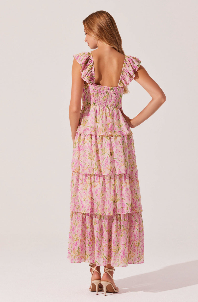 A woman is wearing a long, pink floral dress with tiered ruffles. The dress has a square neckline and ruffled shoulder straps. The fabric features a light pink background with green and darker pink floral patterns. She is standing with her back to the camera, with one hand resting on her hip, and is wearing beige high-heeled shoes.