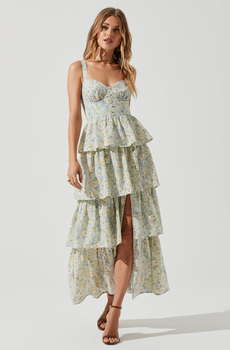 A woman is wearing a sleeveless, multi-tiered dress with puffed bodice and square neckline. The dress features a light-colored, intricate floral and abstract pattern throughout. She pairs the dress with open-toed, strappy high heels.