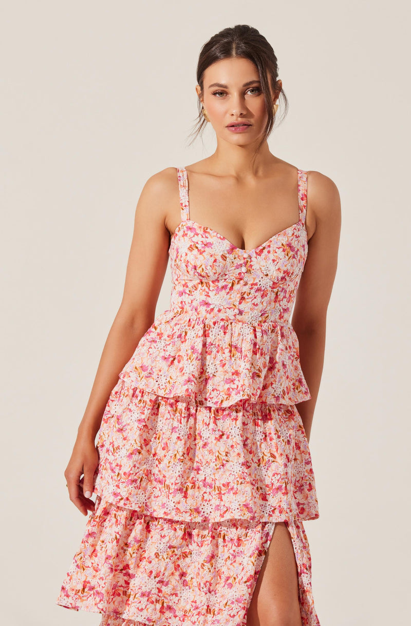 A woman wearing a pink and white floral sleeveless dress with adjustable straps, a fitted bodice, a tiered ruffled skirt, and a slit on the side.