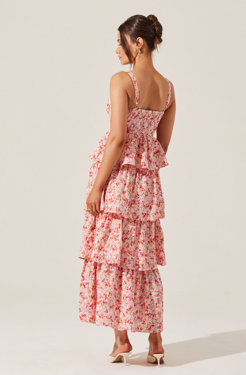 A woman is wearing a sleeveless, floral maxi dress with multiple ruffled layers. The dress features thin shoulder straps and a fitted bodice, with a pattern of pink, red, and orange flowers on a light background. She is also wearing a pair of white heeled shoes and gold earrings. Her hair is styled in a neat bun.