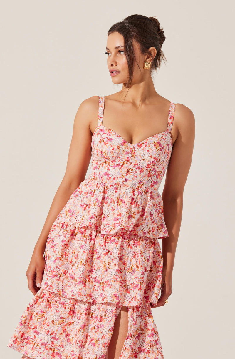 A woman is wearing a sleeveless, floral print dress with a fitted bodice, adjustable spaghetti straps, and layered ruffles on the skirt. The dress features a mix of pink, orange, white, and purple colors in a small, intricate floral pattern.