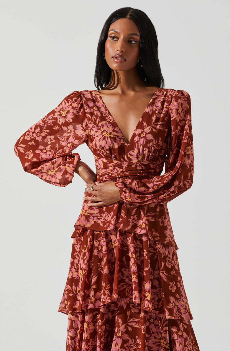 A woman is wearing a long-sleeved, floral-patterned dress with a deep V neckline. The dress has a fitted waist, puffed shoulders, and a tiered, ruffled skirt. The fabric features a reddish-brown background with pink and yellow flowers and green leaves.