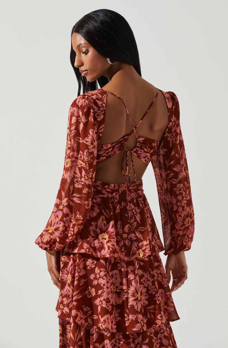 A woman is wearing a matching set of a floral print top and skirt. The long-sleeved top features a back cutout with tie straps, and the fabric has a reddish-brown background with pink and orange floral patterns. The skirt is high-waisted with multiple layered ruffles, matching the floral design and colors of the top.