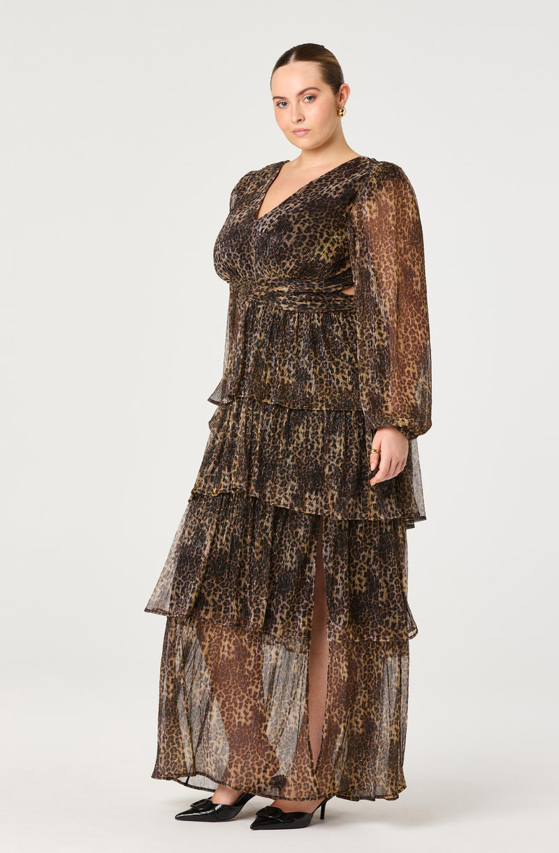 A woman is wearing a long, flowing dress with a leopard print pattern in shades of brown, black, and tan. The dress features long sheer sleeves, a V-neckline, a fitted waist, and a tiered skirt with multiple ruffles.