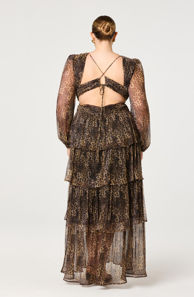 A woman is wearing a long, flowing skirt with multiple tiers, all made from sheer, lightweight fabric featuring a leopard print pattern in shades of brown and black. She is also wearing a matching top with leopard print, long sheer sleeves, and a unique back design with crisscrossed straps and a central tie.