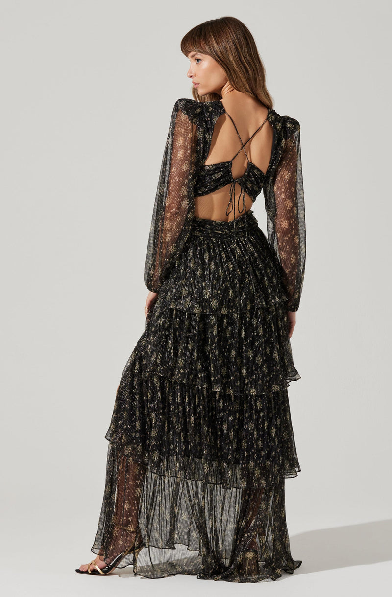 A woman is wearing a black, semi-sheer, long-sleeved top with a floral pattern and puffed shoulders. The top has a tie detail at the back. She is also wearing a high-waisted, tiered, floor-length black skirt with a floral print, made of a lightweight, sheer fabric. She has on black open-toe footwear with a thin strap.