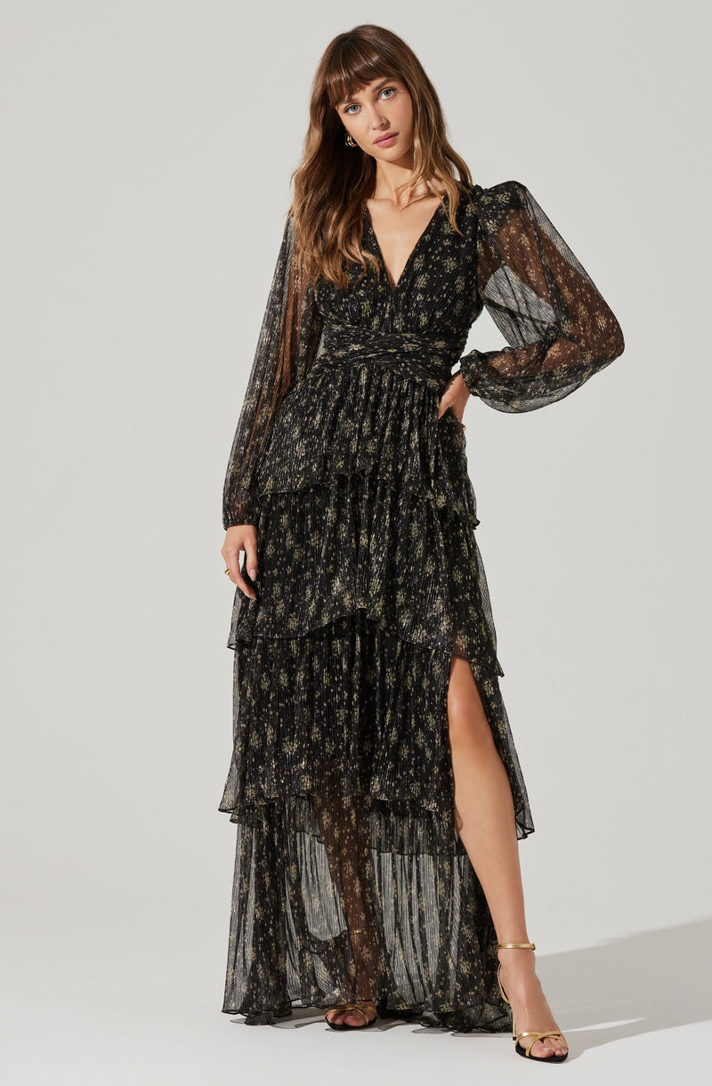 A woman is wearing a long, black dress with a gold floral pattern. The dress has sheer, long puff sleeves with cuffs, a V-neckline, a fitted waist, and a high slit on the left side revealing her leg. The skirt is layered and flows down to the floor.