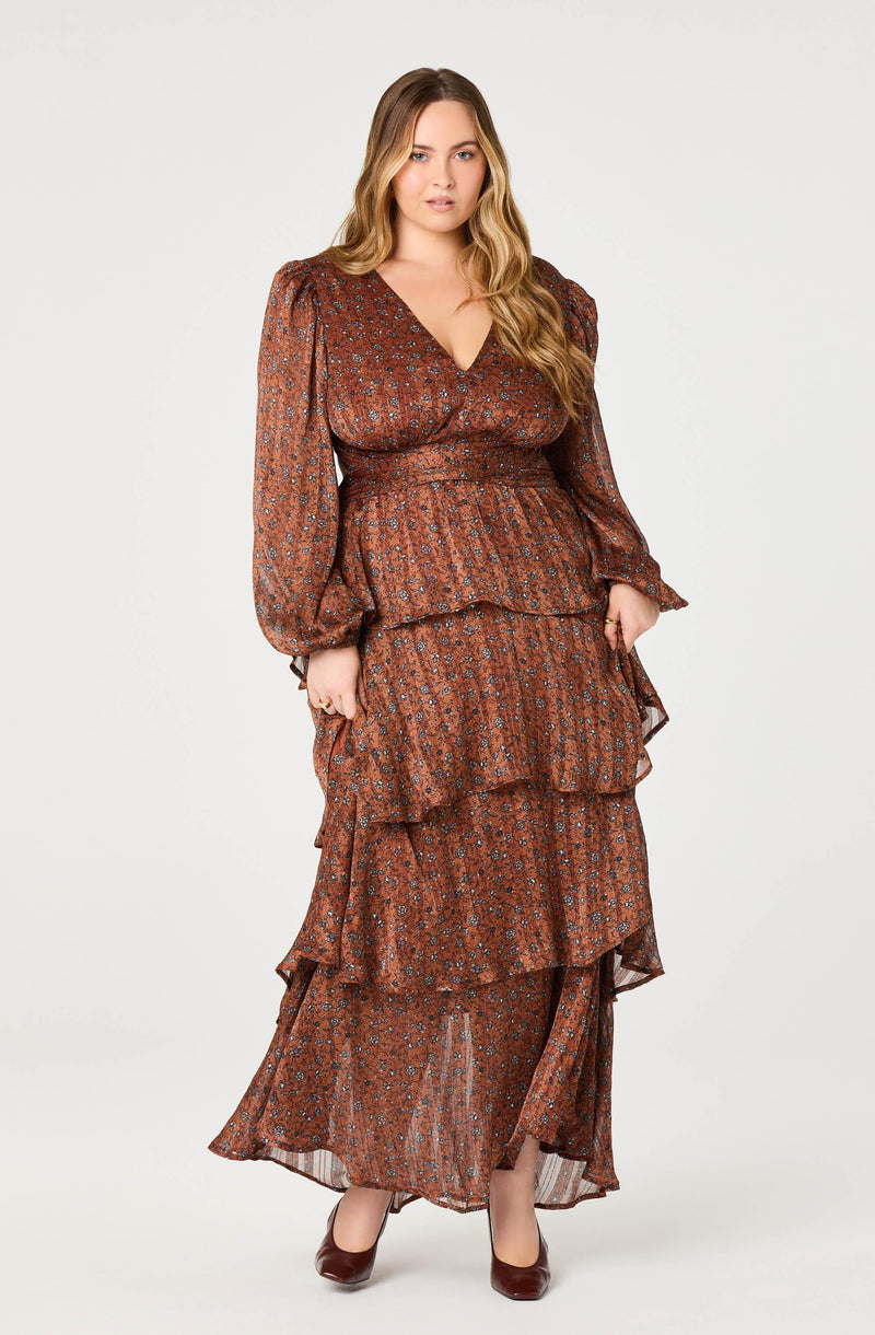 A woman is wearing a long, flowing, brown dress with a small floral pattern. The dress features a V-neckline, long puffed sleeves with elastic cuffs, and multiple layered tiers throughout the skirt. She is also wearing dark brown heels.