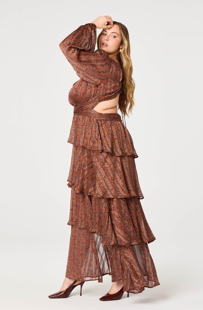 A woman is wearing a coordinated outfit that features a long-sleeved, rust-colored top with a floral pattern. The top has puffed sleeves and a cutout at the side, revealing a portion of her midriff. She is also wearing a matching pair of wide-leg, high-waisted pants with a multi-tiered ruffle design, made from a flowing, semi-sheer fabric with a similar floral pattern. She completes the look with dark brown pointed-toe high heels.